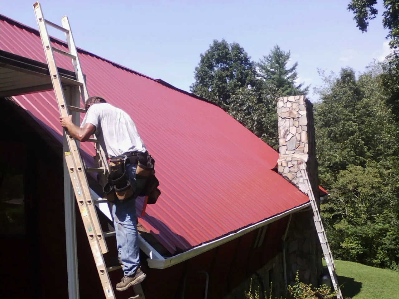 Roofer on a ladder beside a red metal roof and stone chimney — Soffit & Fascia Repair by Roofing Brothers in Foxboro