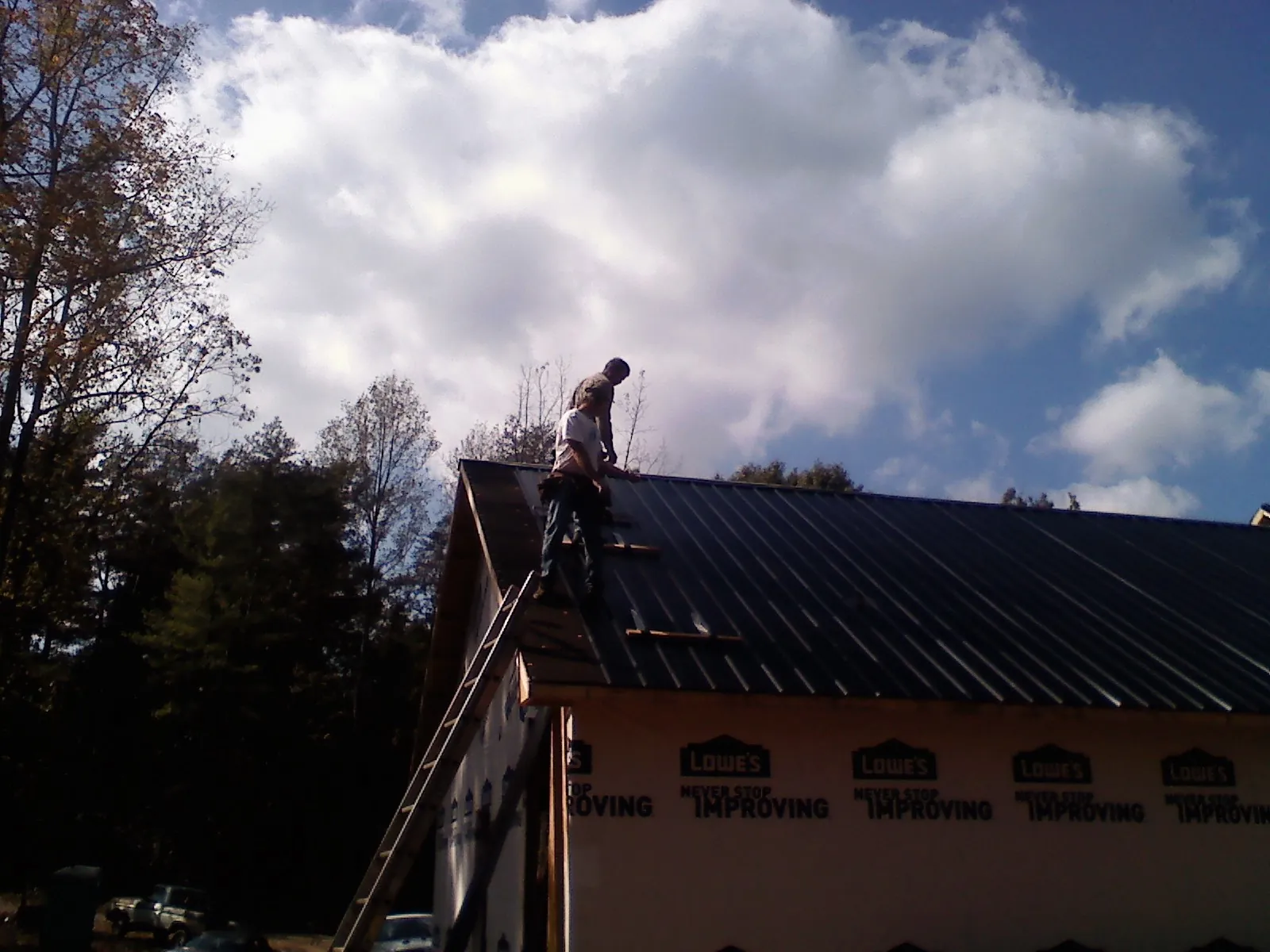 Roofer fastening dark metal panels high against a cloudy sky, from a Roof Inspection job in Foxboro